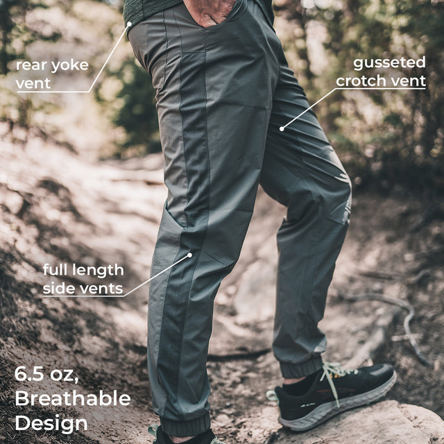 A person outdoors wearing dark gray technical pants with overlaid callouts pointing to rear yoke vent, gusseted crotch vent, full length side vents, and 6.5 oz breathable design.