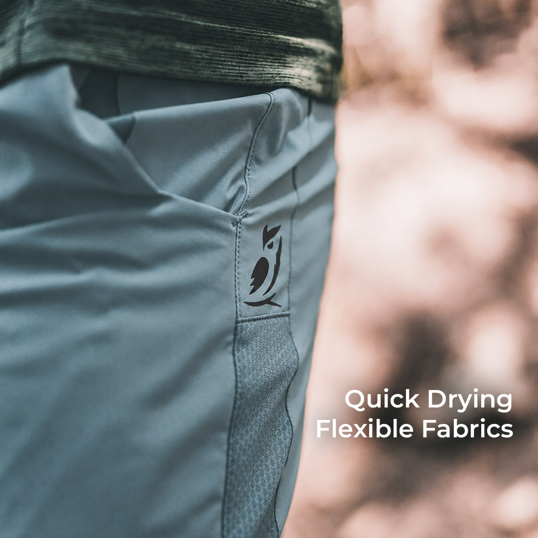 Close-up of a person wearing slate blue athletic joggers with a black logo on the pocket, set against a blurred outdoor background with text reading 'Quick Drying Flexible Fabrics'.