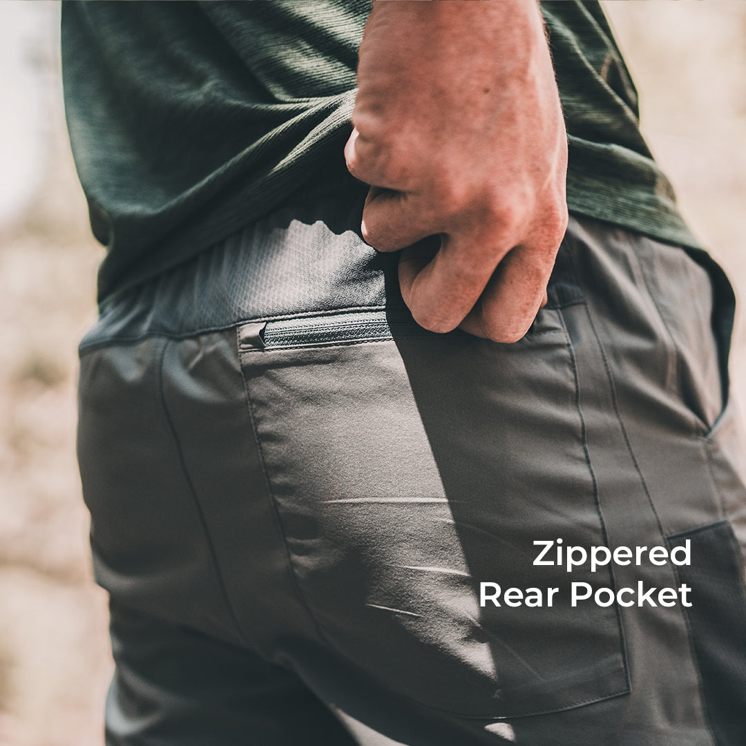 Close-up of a person wearing dark gray zippered rear pocket pants, with a hand reaching into the pocket. White text on the lower right reads 'Zippered Rear Pocket'.