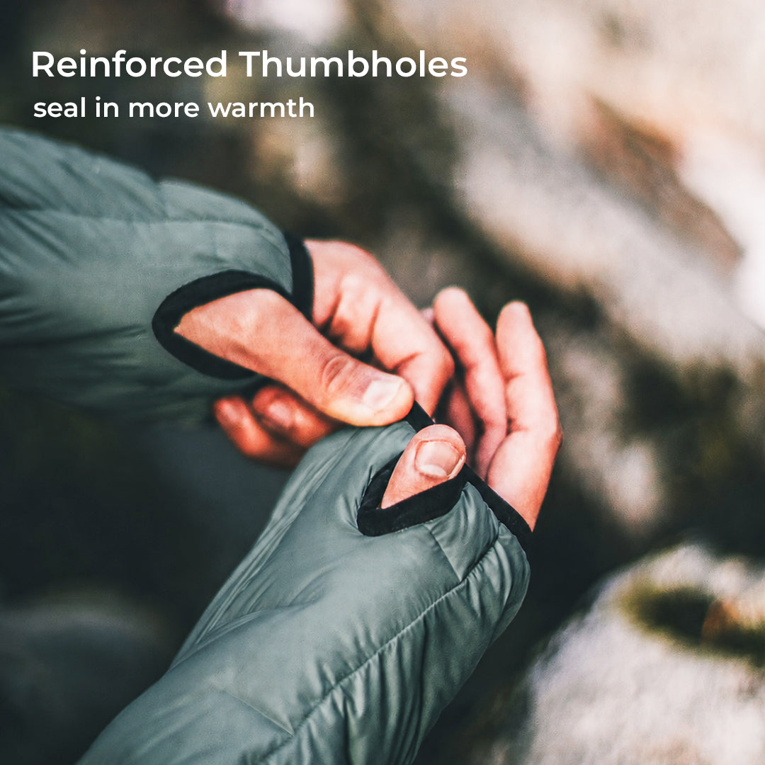 Close-up of a person wearing a dark olive green insulated jacket with thumbholes, adjusting the fit. The text 'Reinforced Thumbholes seal in more warmth' is displayed in the upper left corner against a blurred outdoor rocky background.