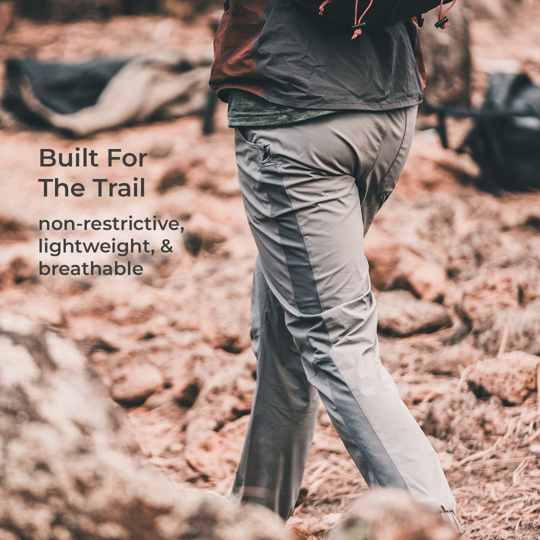 A person wearing light gray hiking pants with dark side panels walks through rocky, reddish-brown outdoor terrain. Text on the left reads, 'Built For The Trail' and 'non-restrictive, lightweight, & breathable.