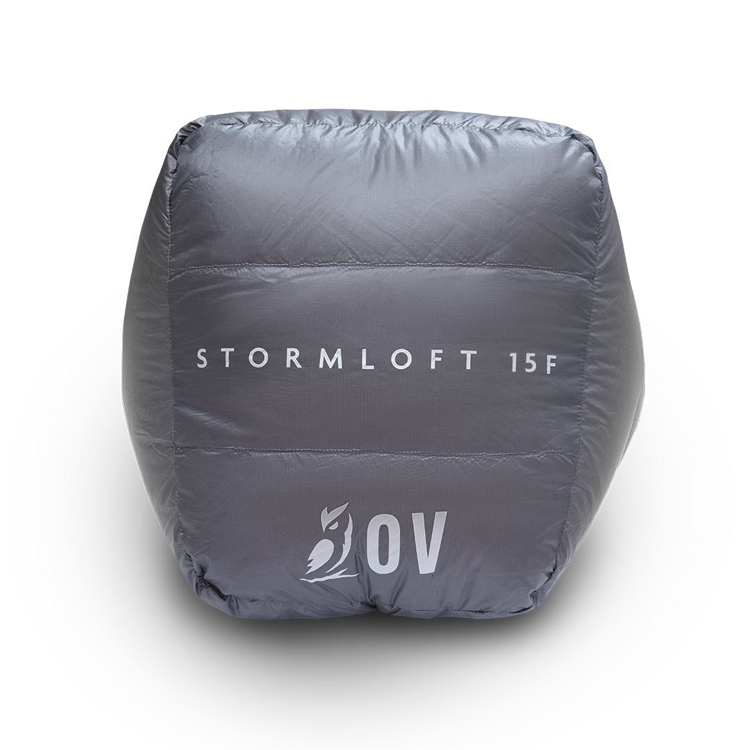 Bottom view of the blue Outdoor Vitals StormLoft 15F topquilt packed inside its compression stuff sack, displaying the printed product name and OV owl logo.