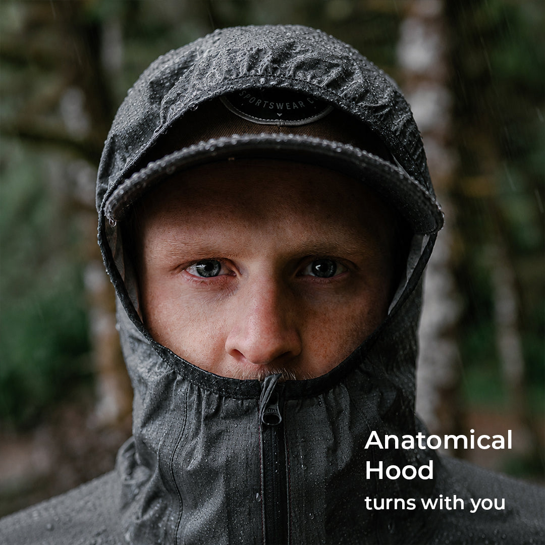 A close-up portrait of a man wearing a waterproof hooded jacket in a blurred outdoor forest setting. The hood is up, covering his face, with the text 'Anatomical Hood turns with you' in the lower right corner.