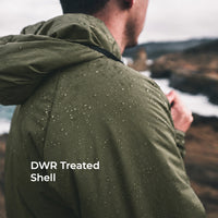 A man viewed from behind wearing an olive green hooded jacket covered in water droplets, with a blurred outdoor coastal landscape in the background. White text on the lower left reads 'DWR Treated Shell'.
