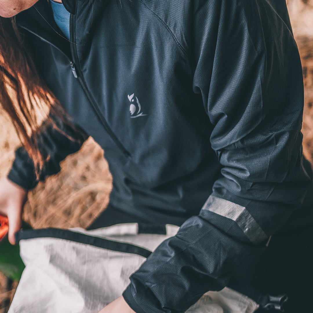 A close-up of a person wearing a synthetic jacket with a small white logo on the chest, sitting outdoors next to a light-colored bag.