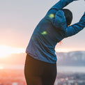 A woman in a blue long-sleeve athletic top and black leggings stretches her arm over her head in an outdoor setting at sunset, with a blurred cityscape and mountains in the background.