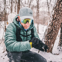 A man in winter gear, including a puffer jacket, black gloves, a gray cap, and mirrored ski goggles, checks his smartwatch while kneeling in the snow next to a tree trunk.