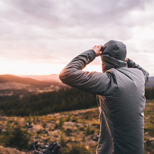 A person wearing a gray hooded jacket stands outdoors on a mountain landscape at sunset, looking out over the scenery as they raise their hood.