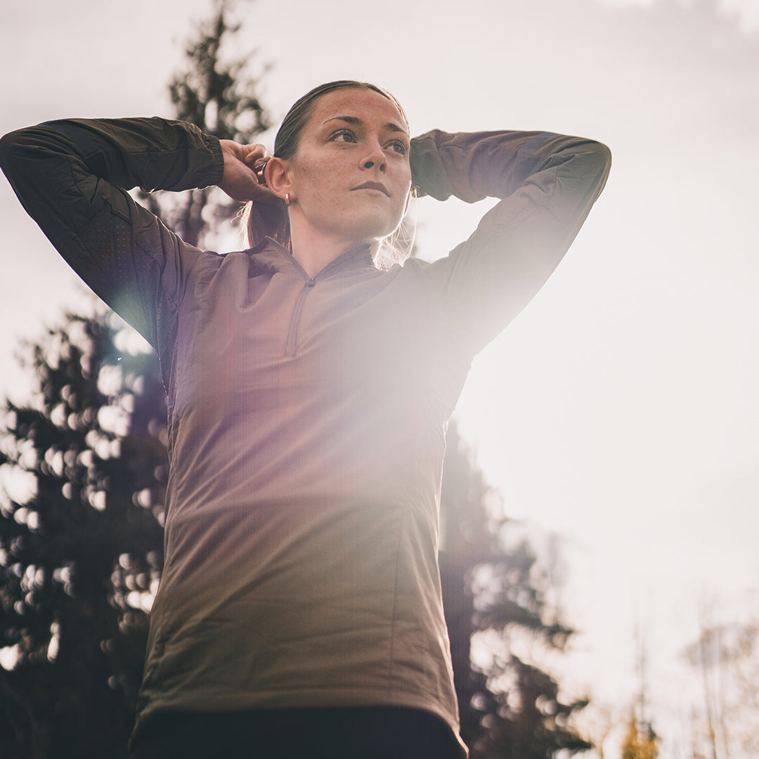 A woman outdoors with her hands behind her head adjusting her ponytail, wearing a light brown quarter-zip athletic pullover and dark pants, set against a bright, sunlit sky and blurred trees.