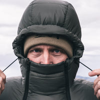 A close-up of a man wearing a dark  puffer jacket with an integrated hood and face warmer, paired with a tan beanie, against an overcast sky.