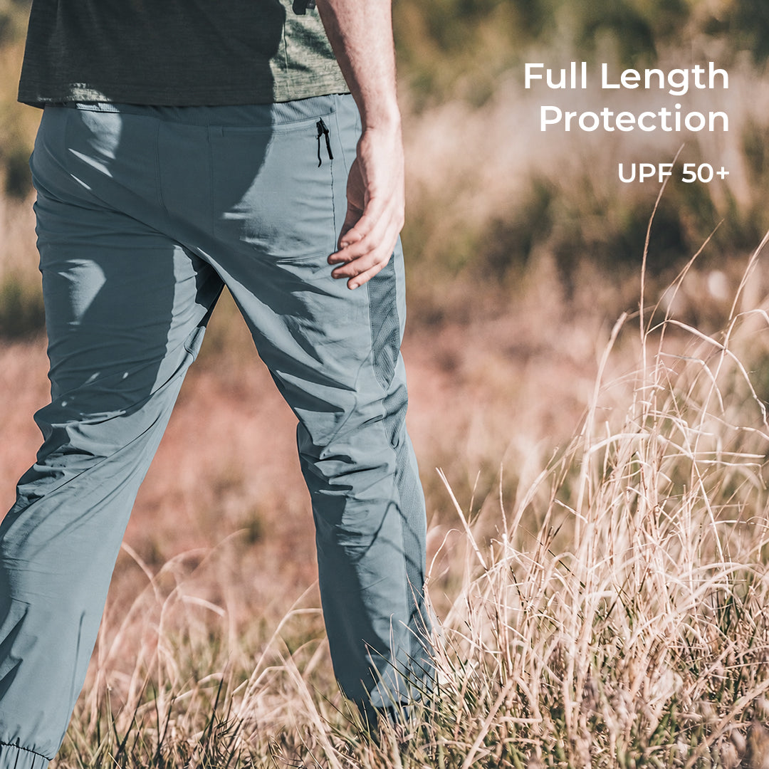 A person outdoors in tall dry grass wearing light blue full-length hiking pants, with text in the upper right reading 'Full Length Protection' and 'UPF 50+'.