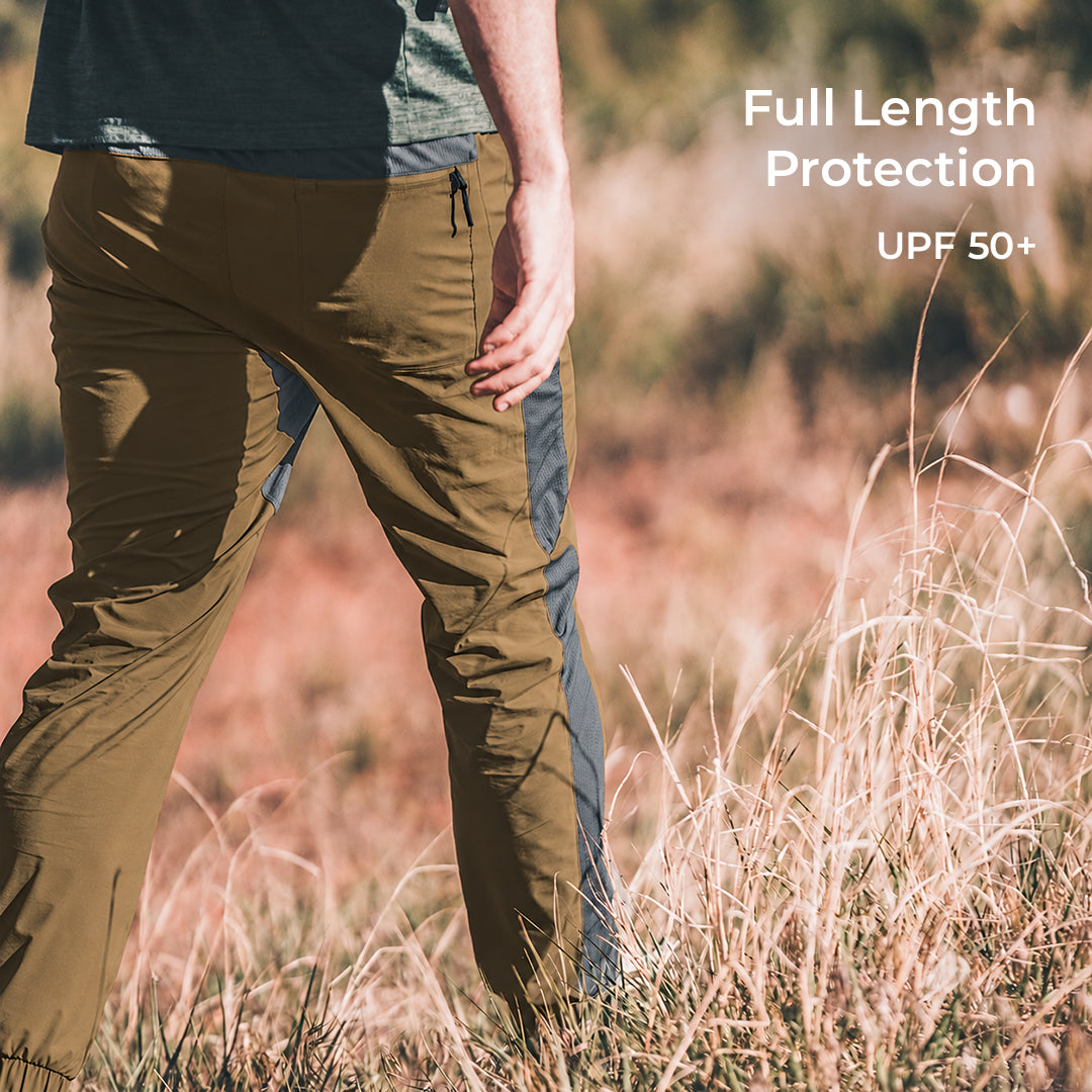 A person outdoors in a grassy field wearing brown full-length hiking pants with dark gray side panels. White text in the upper right reads 'Full Length Protection UPF 50+'.
