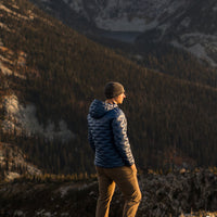 Person wearing a blue Outdoor Vitals insulated jacket and knit beanie, standing on a mountain ridge at sunset and looking out over a forested valley.
