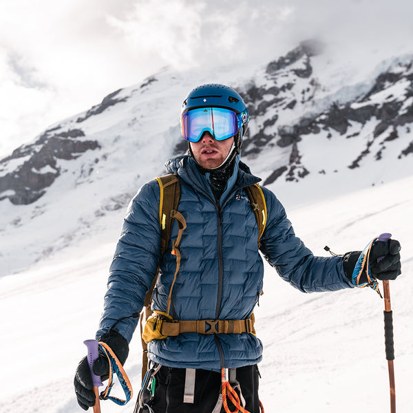 Mountaineer wearing a Outdoor Vitals insulated jacket, helmet, and ski goggles, holding trekking poles while standing on a snowy mountain slope with rugged peaks in the background.