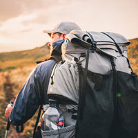 Back view of a hiker wearing the Outdoor Vitals Shadowlight backpack, carrying a rolled sleeping pad secured on top, hiking at golden hour with water bottle visible in side pocket.