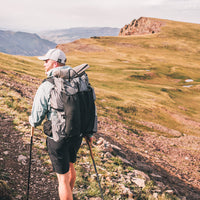 Back view of a hiker in shorts wearing the Outdoor Vitals Shadowlight backpack with a sleeping pad attached, ascending a sunlit alpine ridge.