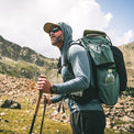 A man hiking in a rocky mountain landscape wearing a long-sleeve hooded shirt, shorts, sunglasses, a tan cap, and carrying a large hiking backpack with a water bottle pocket.