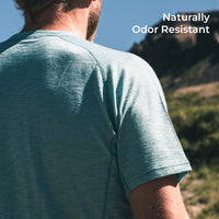 A man wearing a heathered short-sleeve athletic t-shirt, shown from the back in an outdoor mountain setting. White text in the top right corner reads 'Naturally Odor Resistant'.