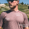 A man wearing a lightweight wool short-sleeve athletic shirt outdoors on a grassy hillside, with white text callouts pointing to raglan sleeves, flatlock seams, stretchy fabric, and 'Designed For Comfort'.