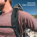 A close-up outdoor shot of a person wearing a backpack over a long-sleeve shirt, with blue sky and a rocky hillside in the background. White text in the top right reads 'Naturally Odor Resistant'.