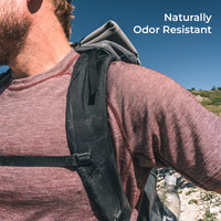 A close-up outdoor shot of a person wearing a backpack over a long-sleeve shirt, with blue sky and a rocky hillside in the background. White text in the top right reads 'Naturally Odor Resistant'.