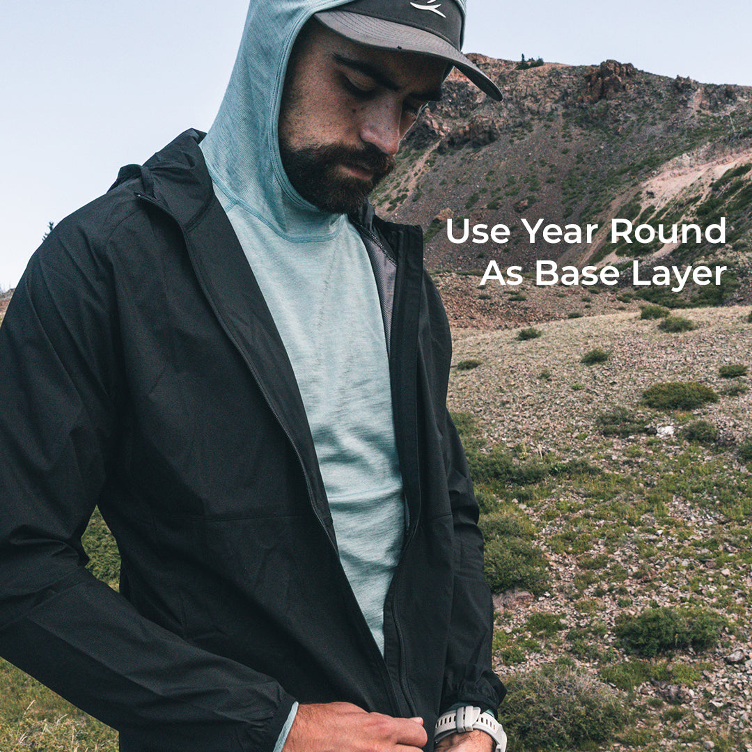 A man wearing a black lightweight zip-front jacket over a light hooded base layer, standing outdoors in a rocky, dry mountain landscape. White text on the right reads 'Use Year Round As Base Layer'.