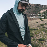 A man wearing a black lightweight zip-front jacket over a light hooded base layer, standing outdoors in a rocky, dry mountain landscape. White text on the right reads 'Use Year Round As Base Layer'.