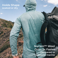 A man wearing a light blue long-sleeve hooded shirt is shown from behind in an outdoor mountain setting, carrying a black backpack. Text overlays highlight the shirt's shape-holding and drying performance using NuYarn Wool.