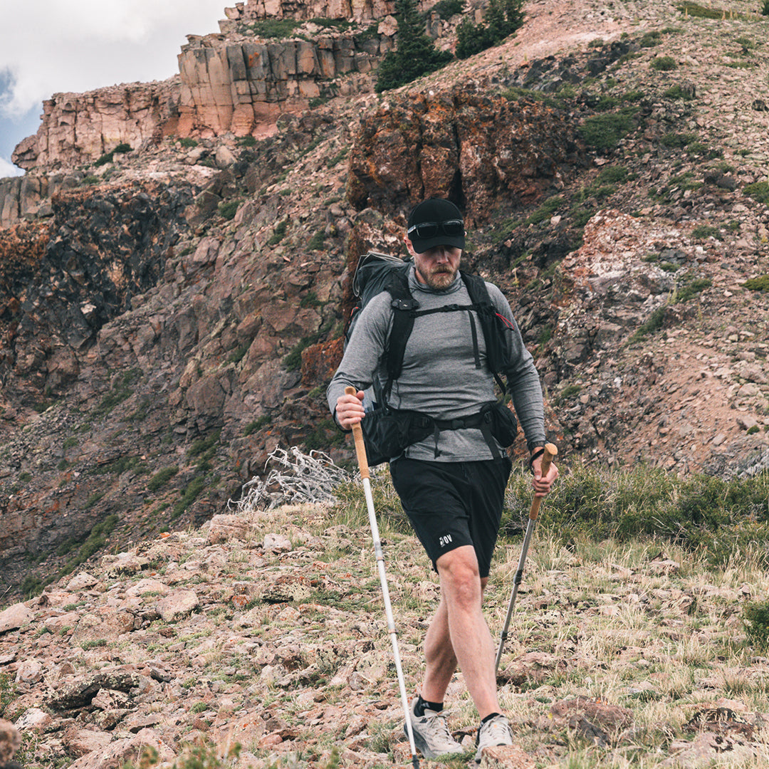 A man hiking on a rocky mountain slope using trekking poles, wearing a long-sleeve top, shorts, and a backpack.