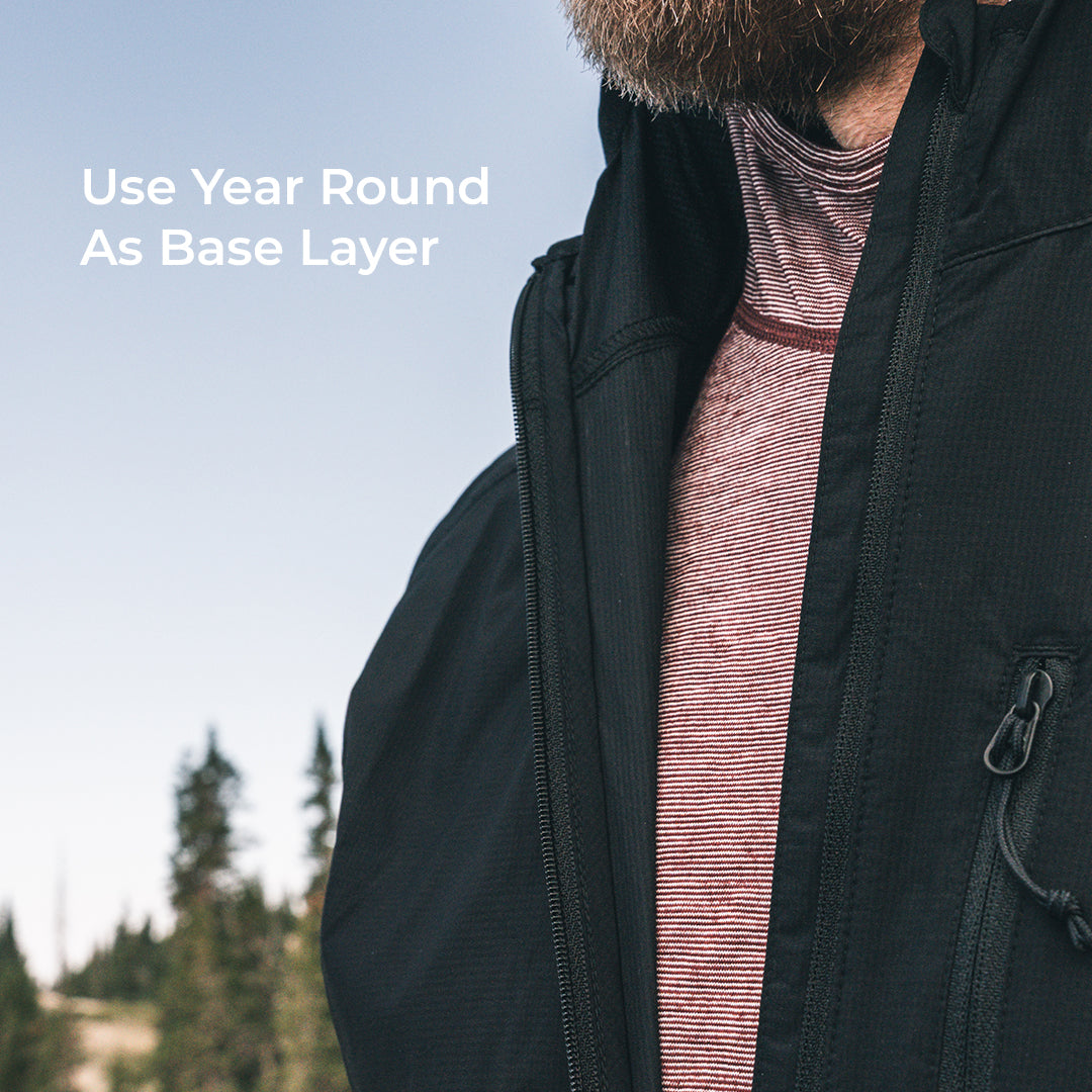 Close-up of a man wearing a black insulated jacket over a wool base layer outdoors, with text on the left reading 'Use Year Round As Base Layer'.