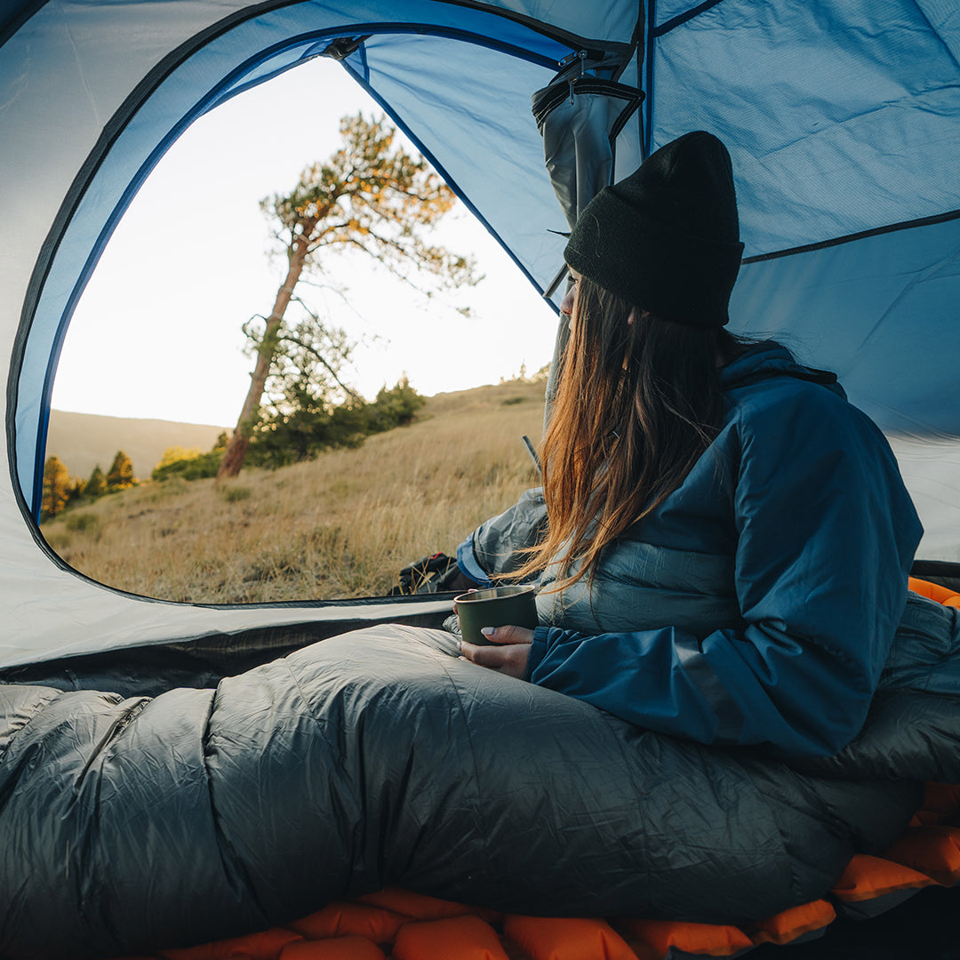 Camper sitting inside a tent in a down top quilt and jacket, holding a cup and looking out at a grassy hillside with trees during sunrise or sunset.