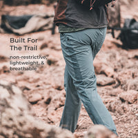 A person outdoors on a rocky trail wearing light blue-gray hiking pants and a dark jacket. Text on the left reads, 'Built For The Trail non-restrictive, lightweight, & breathable.