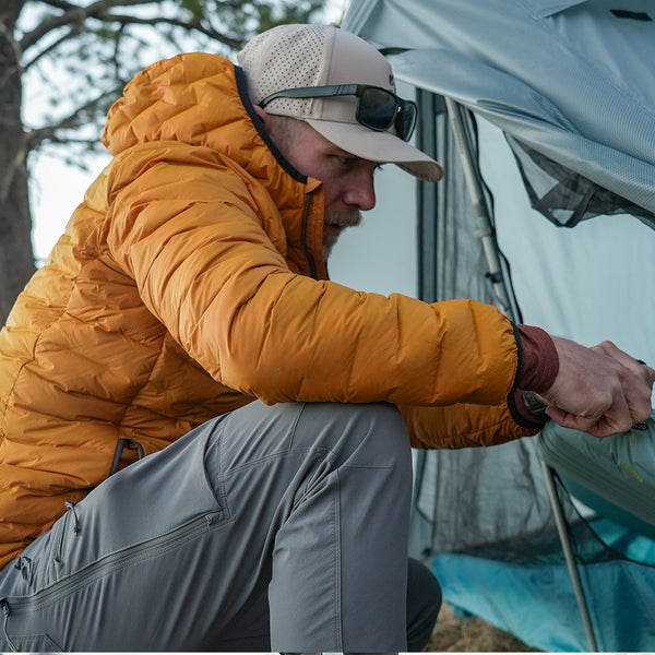 Man wearing insulated down jacket adjusting tent zipper during campsite setup.