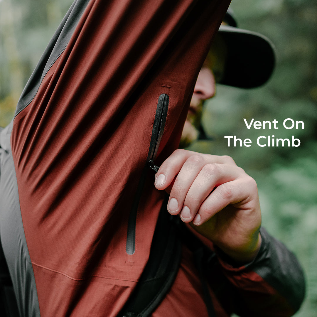 A close-up outdoor shot of a person wearing a ultralight rain jacket, unzipping the underarm vents. The text 'Vent On The Climb' is displayed on the right side against a blurred green forest background.