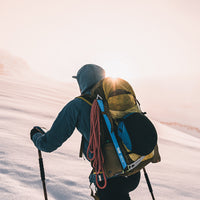 A skier viewed from behind on a snowy mountain slope, wearing a blue jacket and a large yellow and black backpack with a red rope and climbing gear attached. The sun is low on the horizon, creating a bright glow.