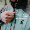 Close-up of a person wearing a waterproof jacket, pulling the zipper with a hand. Water droplets are visible on the jacket, and white text reads 'YKK AquaGuard Zippers everything stays dry'.